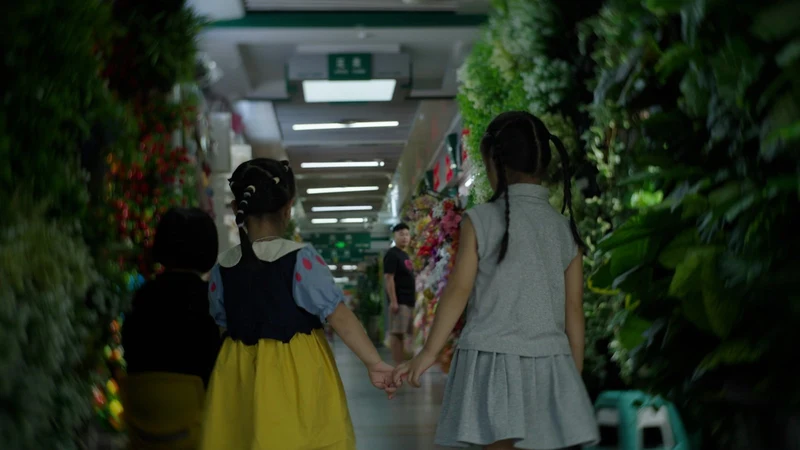 Two girls hold hands as they walk through a market aisle lined with artificial greenery.