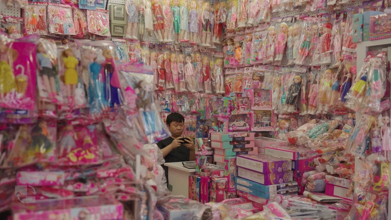 A merchant sits among pink boxes of dolls in a wholesale toy stall.