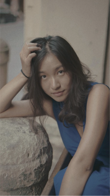 Portrait of Elena Lecun Xiang in a blue dress, leaning on a stone wall.